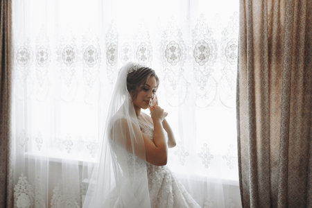 Portrait of the bride. The bride is dressed in an elegant dress, posing by the window wearing earrings. Tiara and jewelry. Open bust. Morning of the brideの写真素材