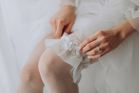 the bride with a French manicure holds a white garter in her hands. Close-up photo. The bride puts on a wedding garter and prepares for the weddingの写真素材