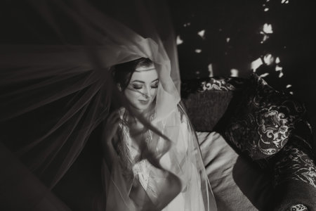Morning of the bride before the wedding. Beautiful young woman under a veil in a white robe on a dark background. Natural beauty and professional makeup. Black and white photoの写真素材