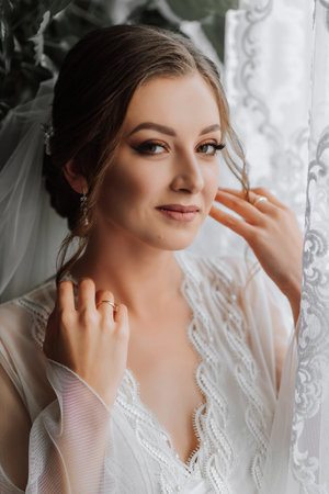 Morning of the bride before the wedding. A beautiful young woman in a white robe near the window. Natural beauty and professional makeup. Portrait of the brideの写真素材