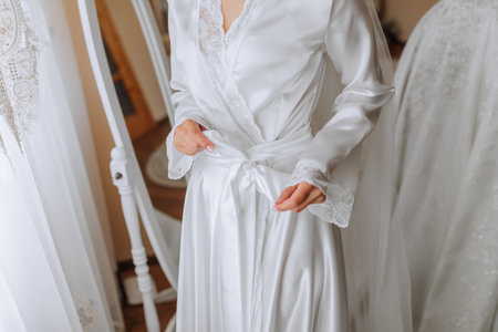 Happy dark-haired brunette bride in a satin robe and professional make-up standing in her room near the mirror and getting ready for her wedding dayの写真素材