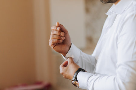 A young groom is buttoning up a white shirt while looking at the camera. The man is wearing a shirt. The groom is preparing for the wedding.の写真素材