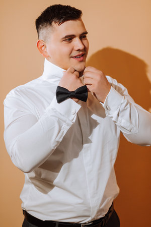 a young smiling man in a white shirt puts on a bow tie. The groom with a short hairstyle on an...
