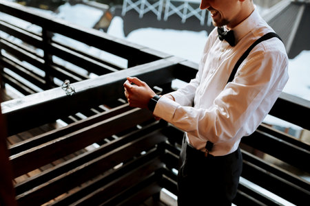 a young man wearing suspenders, a white shirt and a black bow tie puts on his watch. A wedding in the mountains in winterの写真素材