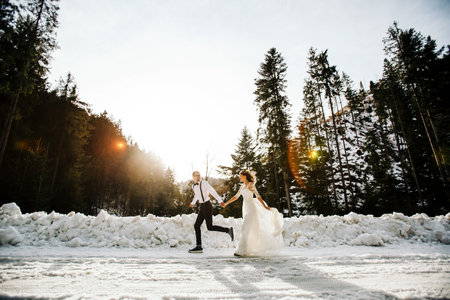 The bride and groom are running along a snowy road against the background of a pine forest and...