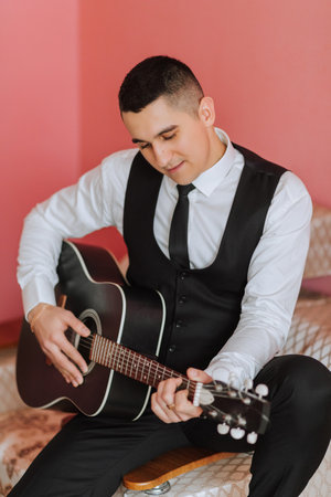 The groom sings songs to the guitar in the morning in his room. Groom in a black suit. Happy man....