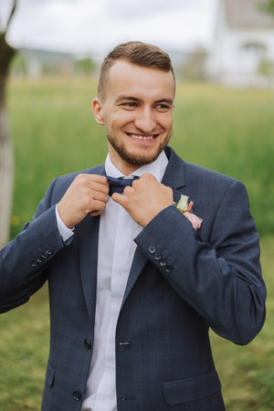 The groom in a black suit adjusts his bow tie, poses against the background of a green tree. Wedding...