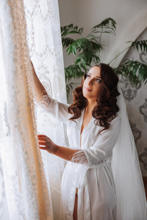 A happy bride is preparing for her luxurious wedding in a hotel room, with a wedding dress on a mannequin nearby. Portrait of a woman with fashionable hair, makeup and a smile in a dressing gown.の写真素材