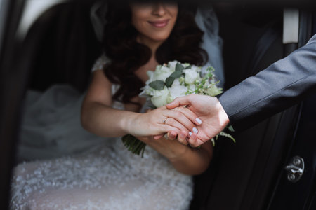 A beautiful bride, sitting in a car, gives her hand to her husband. A beautiful bride with a bouquet of flowers in her hands is sitting in a stylish expensive car.の写真素材