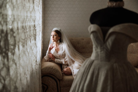 A beautiful bride with a long veil in her room, wearing a robe. Wedding dress on a mannequin. The bride in the morning before the wedding ceremony.の写真素材