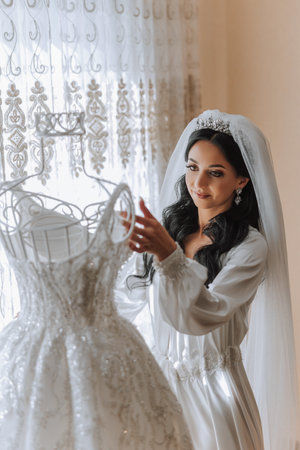 A beautiful bride with a long veil in her room, wearing a robe. Wedding dress on a mannequin. The bride in the morning before the wedding ceremony.の写真素材