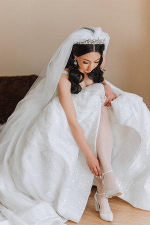 Young bride in beautiful wedding dress putting on shoes indoors. Bride dresses shoes before the wedding ceremony. Detail of bride putting on high heeled sandal wedding shoes. Wedding bride shoes.の写真素材
