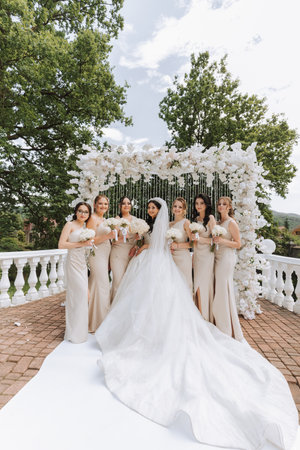 A brunette bride and her bridesmaids in matching cream dresses stand with bouquets near the ceremonial arch. A long train on a wedding dress. Wedding in natureの写真素材