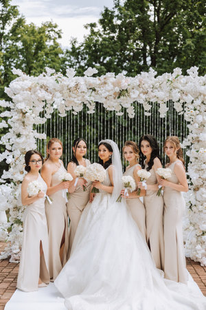 A brunette bride and her bridesmaids in matching cream dresses stand and rejoice with bouquets of flowers near the ceremonial arch. Wedding in natureの写真素材