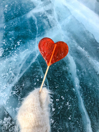 A red lollipop in the shape of a heart holds a hand in a mitten against the background of ice.の写真素材