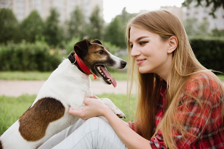 young pretty girl in red tartan shirt and jeans sit at the lawn near high buildings and kiss her dog. Love and friendship concept. Profile view, close up portrait.の写真素材