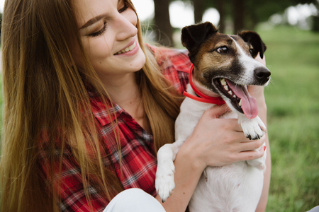 young pretty girl in red tartan shirt and jeans sit at the lawn and hold her dog.の写真素材