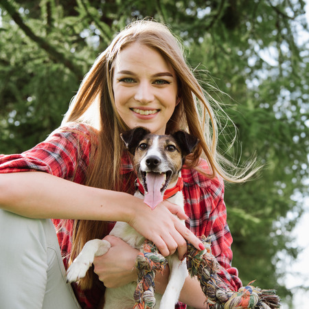 Smiling young pretty girl in red tartan shirt sit at the lawn and embrace her dog.の写真素材