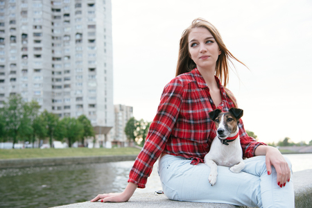 Young woman seat on the embankment near high-rise building. City background. Small fox terrier lies on her lap.の写真素材