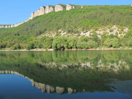 Mirror reflection of a mountain range with steep cliffs and green slopes in a mountain lake. The picturesque landscape of a mountain lake, which reflects the clear blue sky, rocks and mountain slopes, overgrown with green trees and shrubs on a clear summer day, Mangup, Crimeaの写真素材