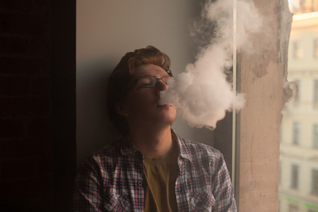 Young man smoking electronic cigarette near window. vaping.の写真素材
