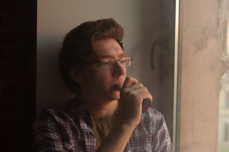 Young man smoking electronic cigarette near window. vaping.の写真素材