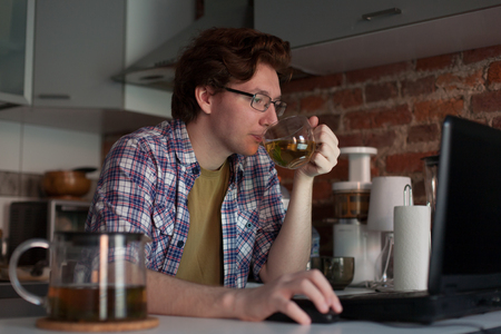 The young man sits at a laptop in the kitchen and drinking tea. Viewing videos.の写真素材