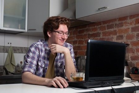 Young man working on laptop. Happy guy is sitting in the kitchen and chatting with his friends.の写真素材