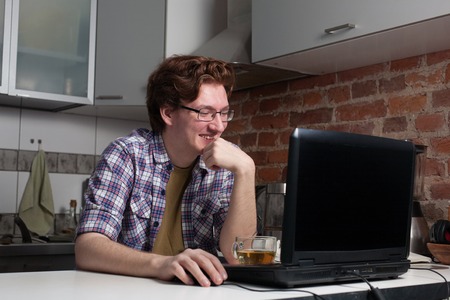 Young man working on laptop. Happy guy is sitting in the kitchen and chatting with his friends.の写真素材