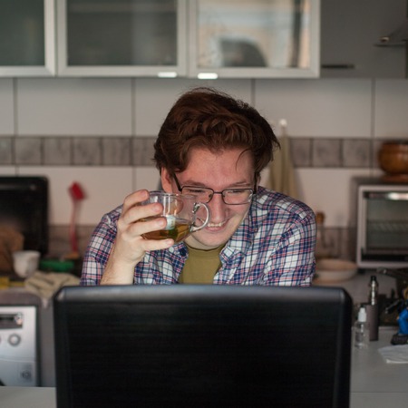 Young man working on laptop. Happy guy is sitting in the kitchen and chatting with his friends.の写真素材