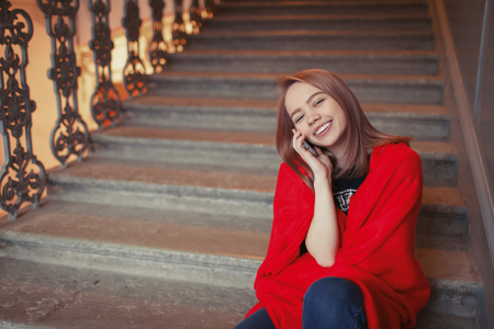 A young blonde girl talking on the phone sitting on the steps, wrapped in a red blanket. She is happy and smiling.の写真素材