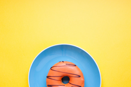 Blue plate with an orange donut in the glaze on a yellow background. Top view with copy space. Minimal concept.の写真素材