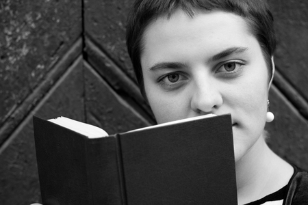 A girl with big eyes and short hair hides behind a book. Close-up portrait. Black and white photo.