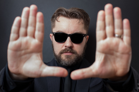 A serious man with a beard and a stylish hairstyle carries out framing with his hands. Directed in a business black suit and sunglasses.の写真素材