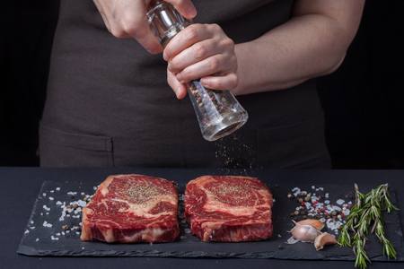 A woman cooks with peppers two fresh raw ribeye steaks from marbled beef on a dark background. Nearby is a mixture of peppers, sea salt, garlic and rosemary.の写真素材