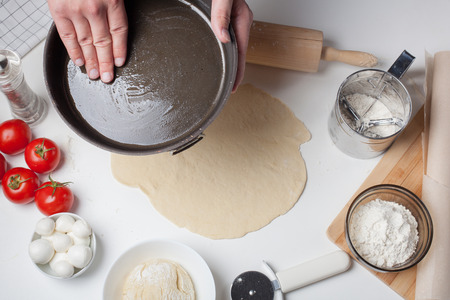 The man-cook spreads olive oil on the pizza mold. On a white table are tomatoes, mozzarella balls, olive oil and flour.の写真素材