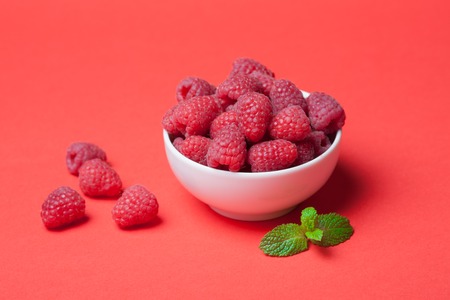 Bowl with fresh raspberries and mint leaves on a red background. Copy space. Minimal concept. hard light.の写真素材