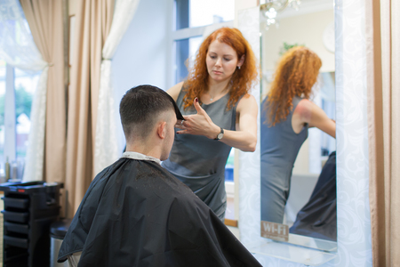 Girl hairdresser with curly red hair cuts young, handsome guy in a beauty salon.の写真素材