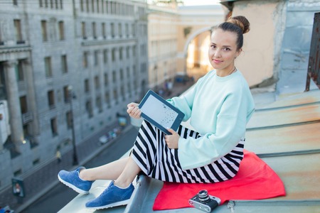 Attractive and stylish girl sits on the roof of the house in the old town and reads the e-book. She sits on a red blanket and lying next to a vintage camera.の写真素材
