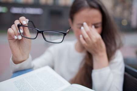 Closeup portrait of attractive female with eyeglasses in hand. Poor young girl has issues with vision. She rubs her nose and eyes out of fatigue. A student tired to study and read books.の写真素材