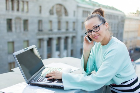 Cute businesswoman in eyeglasses talking on the phone standing on the roof of the house in the old town. Next to it are a laptop, smartphone and paper documents.の写真素材