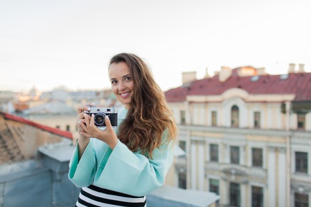 Fashion look, pretty cool young woman model with retro film camera. curly hair outdoors. Stylish girl photographer takes the old city from the roof at sunset.の写真素材