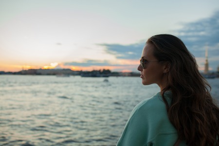 Young and beautiful girl sitting on the embankment of the river. she looks at the sunset and ships passing by. Saint Petersburg, Russia.の写真素材