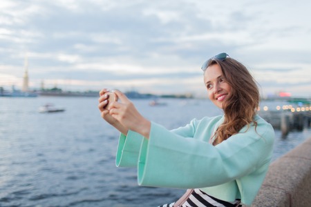 Young and beautiful girl sitting on the embankment of the river. she looks at the sunset and taking a selfie on your phone. Saint Petersburg, Russia.の写真素材