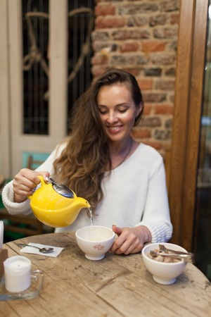 Smiling young girl in the restaurant pours green tea from the yellow teapot in a Cup. Focus on a mug of tea.の写真素材