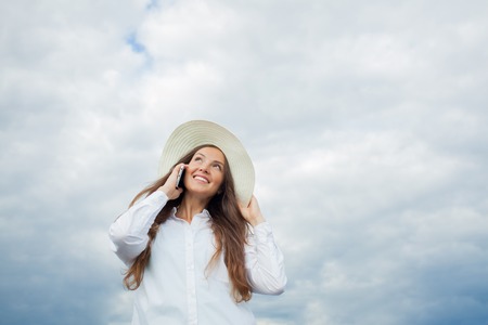 Beautiful smiling girl in a white hat with a wide brim talking on the phone on background of storm clouds.の写真素材