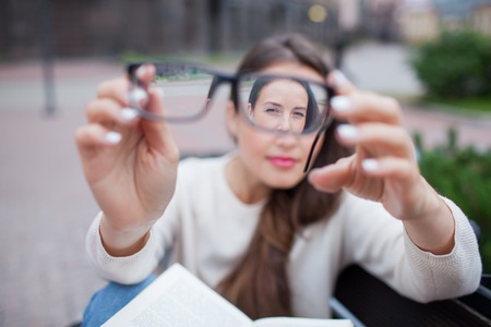 Closeup portrait of young women with glasses. She has eyesight problems and is squinting his eyes a little bit. Beautiful girl is holding eyeglasses right in front of camera with two hand.の写真素材