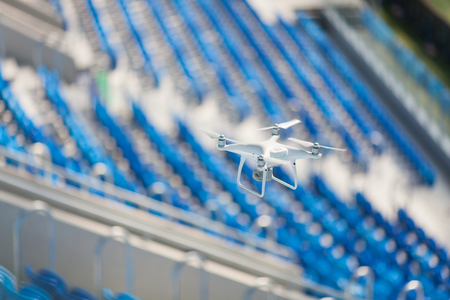 White quadcopter flies over the football stadium and shoots video. The drone on the background of blue seats at the stadium.の写真素材