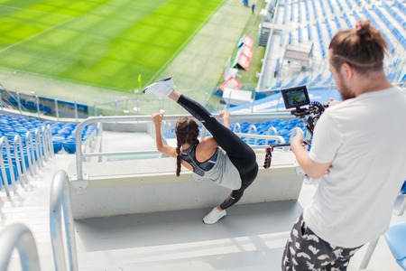 The cameraman shoots on a professional camera, as a sports girl kneads in a stadium against the background of a football field. A sexy woman with a magnificent stretch holds on to a metal railing and raises her leg above her head.の写真素材