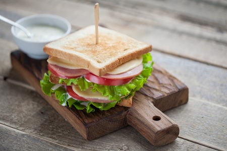 Close-up big sandwich with ham, cheese, tomatoes, salad and white sauce on toasted bread on a rustic wooden table.の写真素材